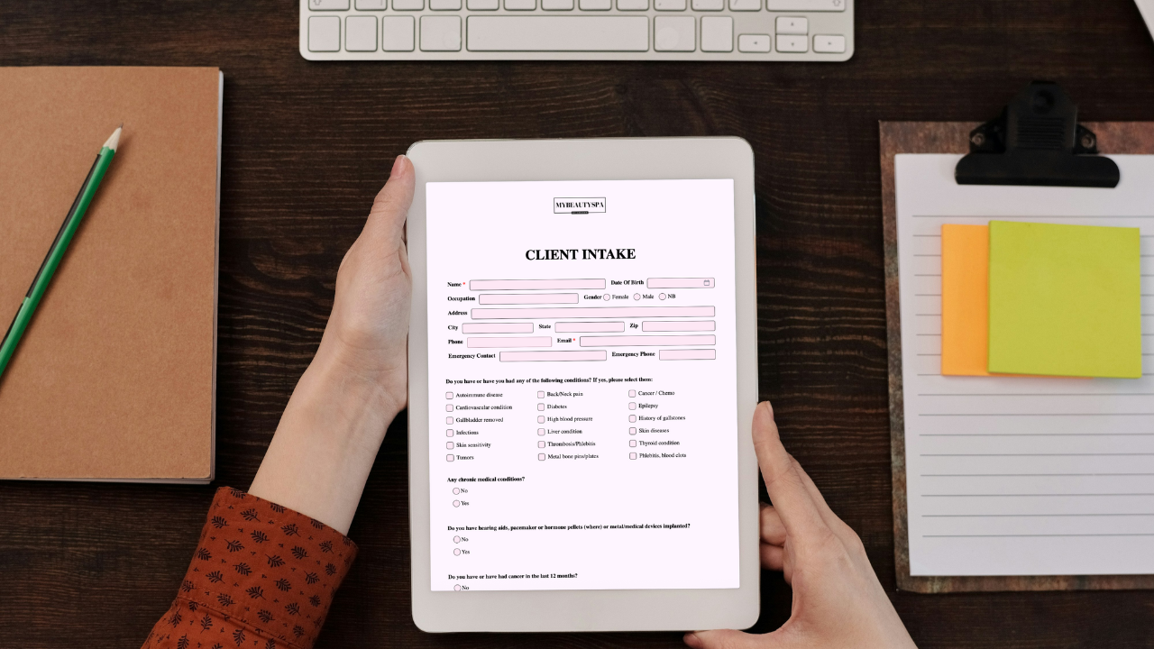 Hands holding a tablet with a Glow Forms client intake form at a salon desk, with keyboard and clipboard nearby.