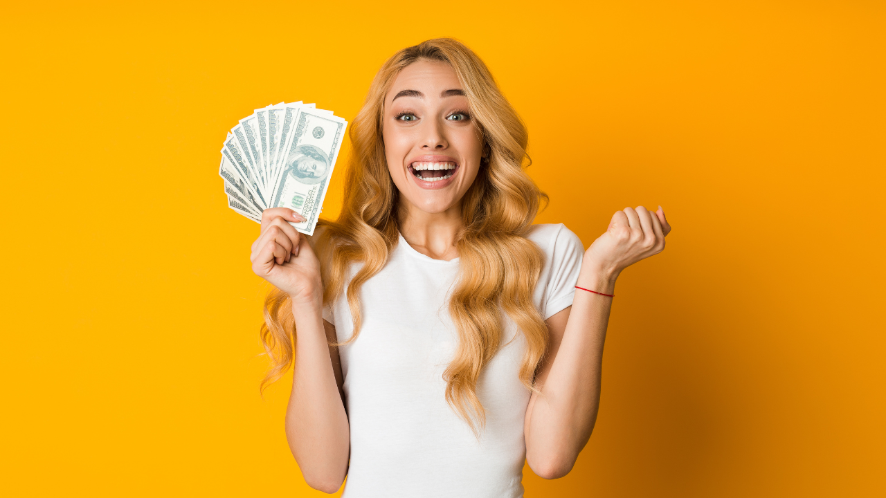 Excited young woman holding a fan of hundred dollar bills against a vibrant orange background, celebrating financial success or business profit