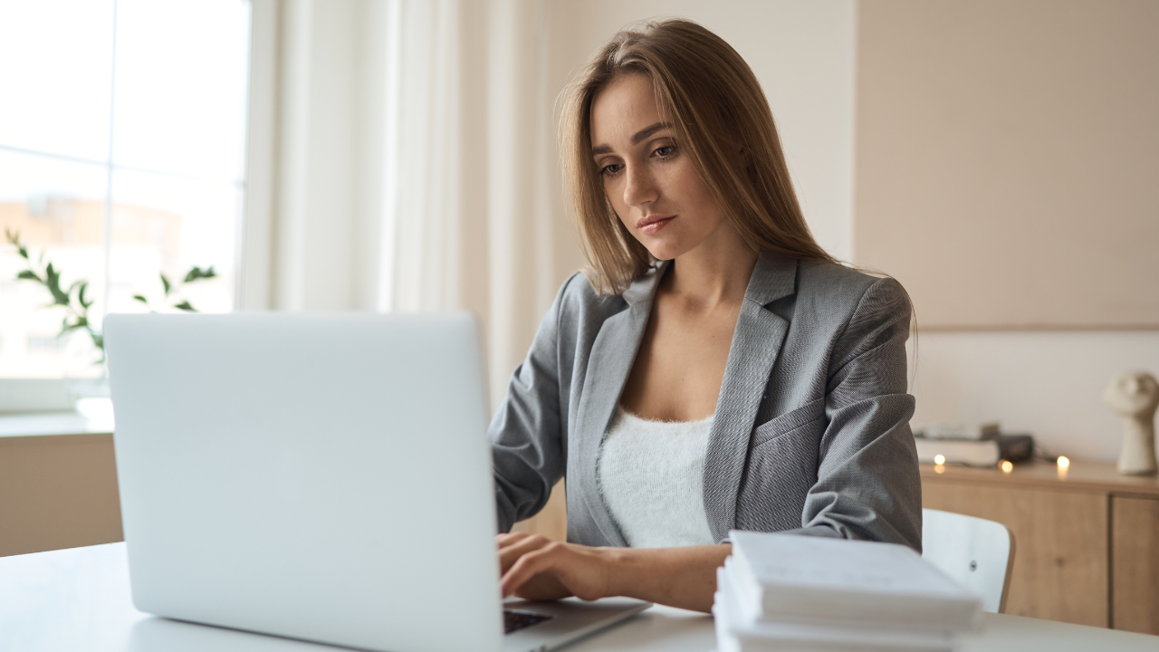 Focused businesswoman in a grey blazer working on a laptop at a modern, softly lit office desk with papers in front of her