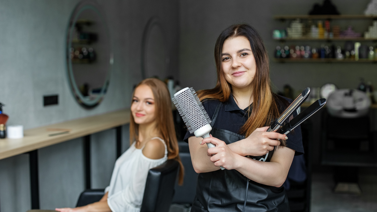 Hairstylist in salon holding a round brush and curling iron, smiling with a client seated in the background before the consultation.
