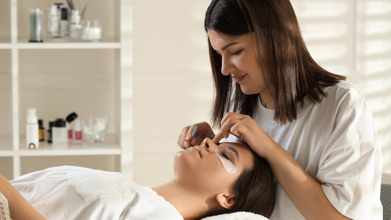 Lash technician applying eyelash extensions with under-eye pads in a bright salon, client relaxed on a treatment bed.