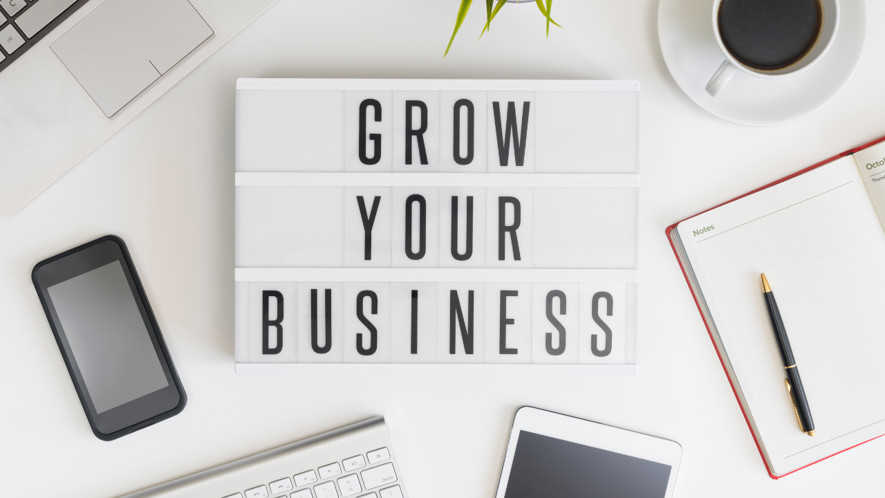 Flat lay with a “Grow Your Business” sign, smartphone, keyboard, tablet, notebook, and coffee—planning beauty business growth.