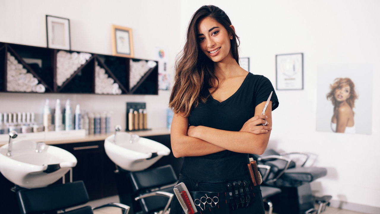 Confident hairstylist stands in her salon with scissors and a tool belt, ready for clients.