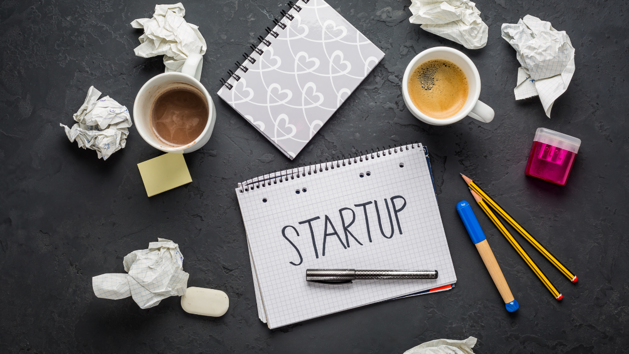 Beauty business startup planning—coffee mugs, crumpled drafts, and a notebook reading “STARTUP” on a workspace.