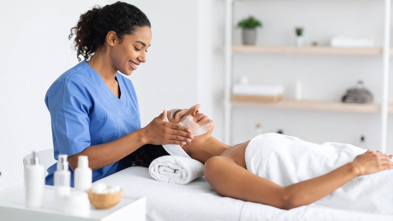 Beauty therapist gently applies a cream mask during a facial treatment in a bright, minimal spa.