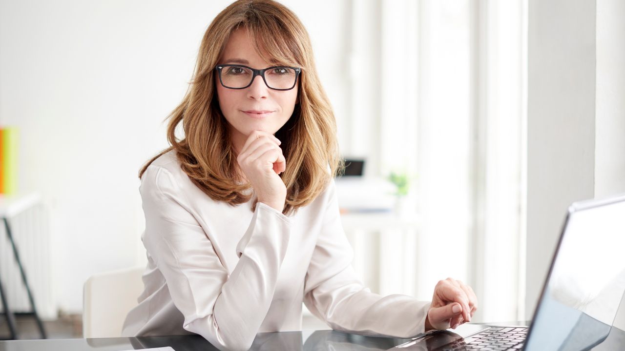 Salon owner reviews business plans on a laptop, looking confident in a light-filled workspace.