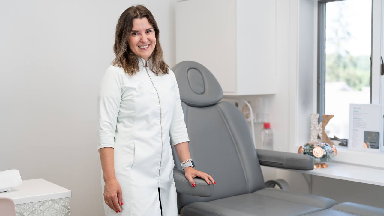 New esthetician in a bright treatment room, ready to welcome clients beside a spa chair.