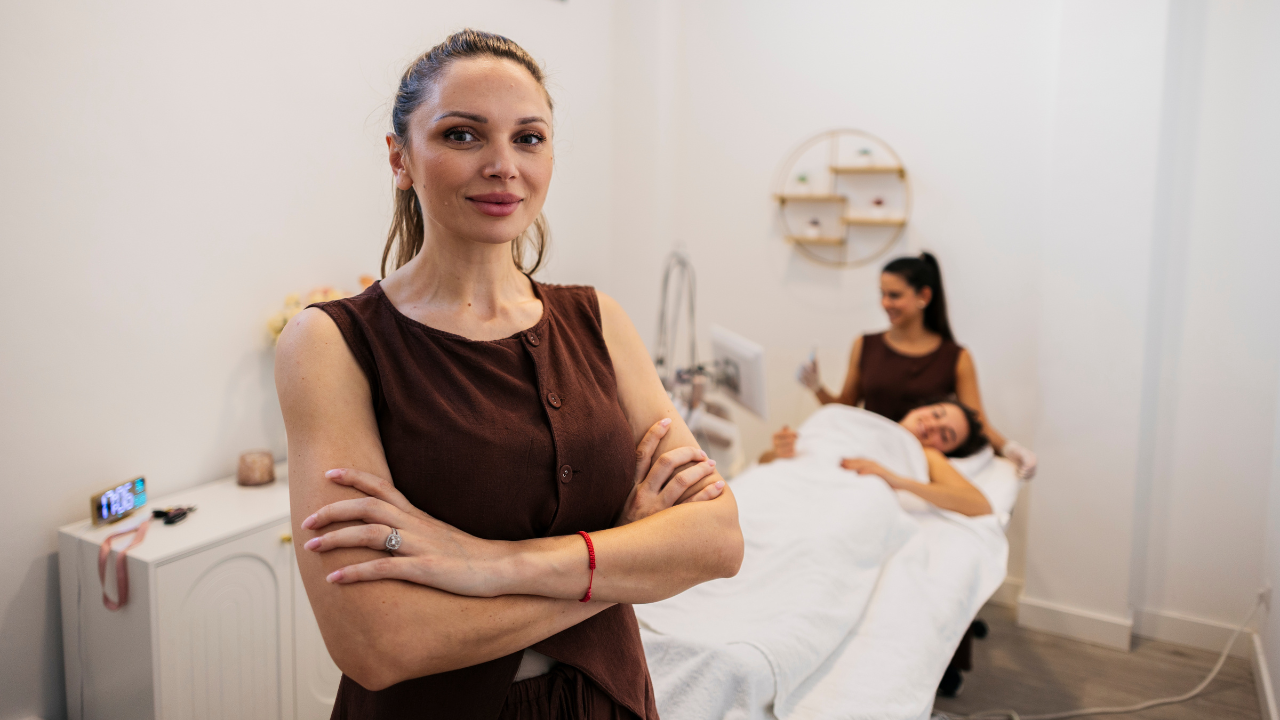 Beauty pro smiles at the camera while a client receives a treatment in the background—calm, welcoming studio.