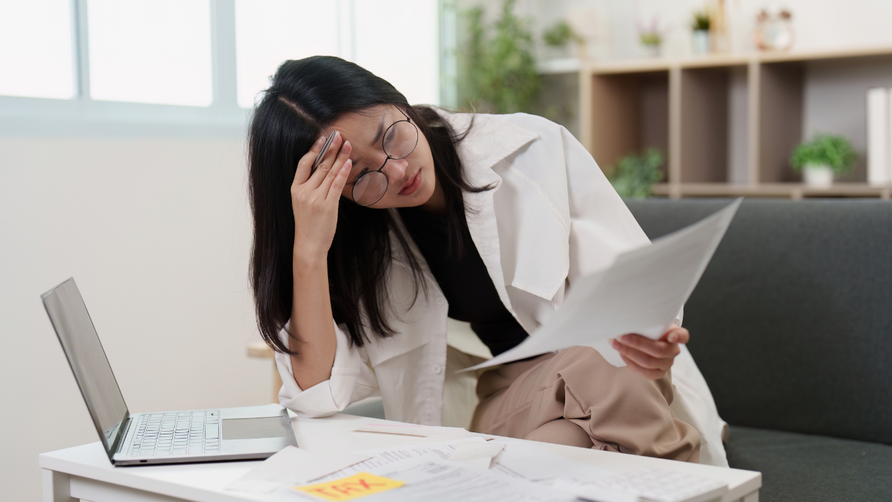 Beauty professional looks worried while checking bills and tax papers at a desk with a laptop.